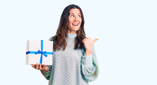 Young beautiful brunette woman holding birthday gift pointing thumb up to the side smiling happy with open mouth