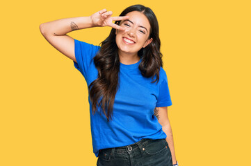 Beautiful brunette young woman wearing casual clothes doing peace symbol with fingers over face,...