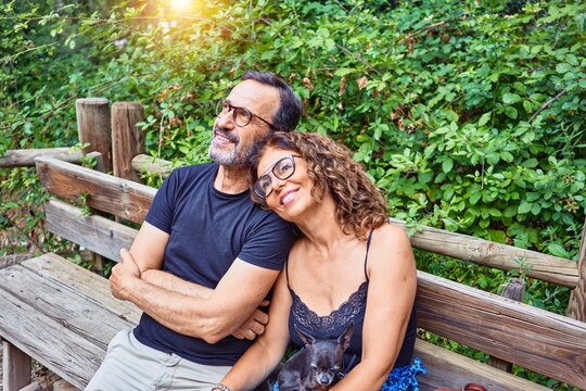 Middle age couple smiling happy looking at the sky. Sitting on the bench with chihuahua dog at park