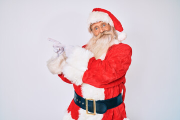 Old senior man with grey hair and long beard wearing santa claus costume with suspenders smiling and looking at the camera pointing with two hands and fingers to the side.