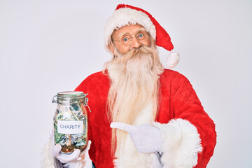 Old senior man wearing santa claus costume holding charity jar smiling happy pointing with hand and finger