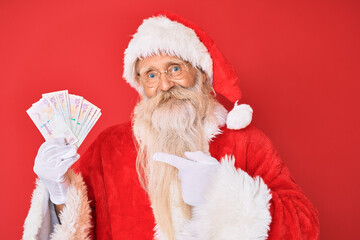 Old senior man with grey hair and long beard wearing santa claus costume holding colombian pesos smiling happy pointing with hand and finger