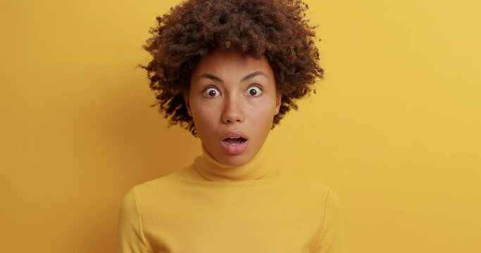 Speechless impressed curly haired young woman stares in disbelief has bugged eyes widely opened mouth stares with shock at camera dressed in casual poloneck poses against yellow studio background