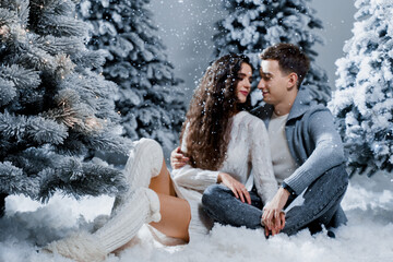 Fototapeta na wymiar New year love story. Couple kisses and hugs. People weared wearing fur headphones, hats, white sweaters. Happy young couple hugs and kiss near christmas trees in winter day.