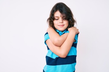 Cute hispanic child with long hair wearing casual clothes hugging oneself happy and positive,...