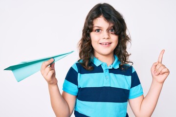 Cute hispanic child with long hair holding paper airplane smiling happy pointing with hand and finger to the side