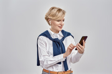 Fototapeta premium Portrait of elegant middle aged caucasian woman wearing business attire and earphones holding, using smartphone while standing isolated over grey background
