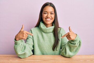 Beautiful hispanic woman wearing casual clothes sitting on the table looking confident with smile...