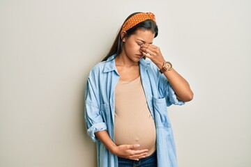 Beautiful hispanic woman expecting a baby, touching pregnant belly tired rubbing nose and eyes...