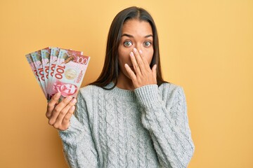 Beautiful hispanic woman holding 100 new zealand dollars banknote covering mouth with hand, shocked and afraid for mistake. surprised expression