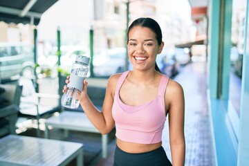 Young beautiful hispanic sporty woman wearing fitness outfit smiling happy and natural at the town drinking fresh water from the bottle