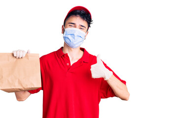 Young hispanic man wearing delivery uniform and medical mask holding paper bag smiling happy and positive, thumb up doing excellent and approval sign