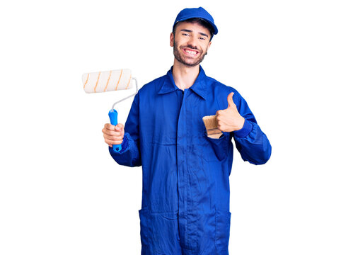 Young hispanic man wearing painter uniform holding roller smiling happy and positive, thumb up doing excellent and approval sign