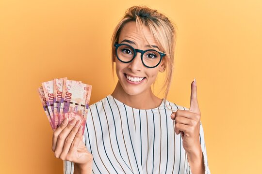 Beautiful blonde woman holding south african 50 rand banknotes smiling with an idea or question pointing finger with happy face, number one