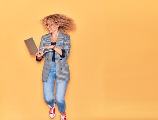 Young beautiful curly businesswoman wearing glasses smiling happy. Jumping with smile on face working using laptop over isolated yellow background.