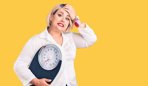 Young Blonde Plus Size Woman Holding Weighing Machine Stressed And Frustrated With Hand On Head, Surprised And Angry Face