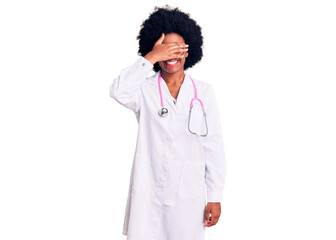 Young african american woman wearing doctor coat and stethoscope smiling and laughing with hand on face covering eyes for surprise. blind concept.