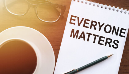 Everyone matters text on paper with a cup of coffee and glasses on a wooden background in sunlight.