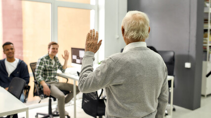 Naklejka na ściany i meble Back view of aged man, senior intern waving, greeting young colleagues at his new job in the modern office