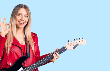 Young beautiful blonde woman playing electric guitar doing ok sign with fingers, smiling friendly gesturing excellent symbol
