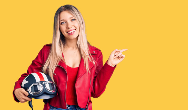 Young beautiful blonde woman holding motorcycle helmet smiling happy pointing with hand and finger to the side