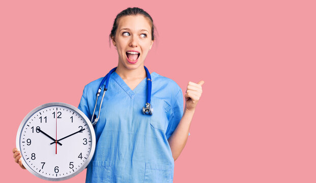 Young beautiful blonde woman wearing doctor uniform and stethoscope pointing thumb up to the side smiling happy with open mouth