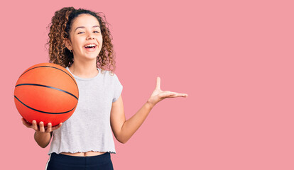 Beautiful kid girl with curly hair holding basketball ball celebrating victory with happy smile and...