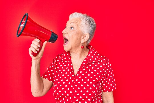 Senior beautiful grey-haired woman screaming using megaphone over isolated red background