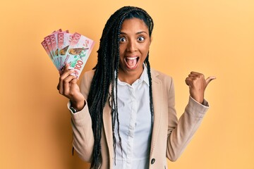 African american woman with braids holding 100 new zealand dollars banknote pointing thumb up to the side smiling happy with open mouth