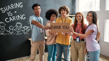 Group of diverse young volunteers smiling at camera, holding card with Thank you lettering while...