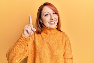 Beautiful redhead woman wearing casual winter sweater over yellow background pointing finger up with successful idea. exited and happy. number one.