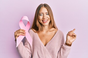 Beautiful young blonde woman holding pink cancer ribbon smiling happy pointing with hand and finger to the side