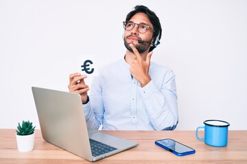 Handsome hispanic man working at the office holding euro banner serious face thinking about question with hand on chin, thoughtful about confusing idea