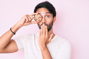 Handsome hispanic man holding cookie covering mouth with hand, shocked and afraid for mistake....