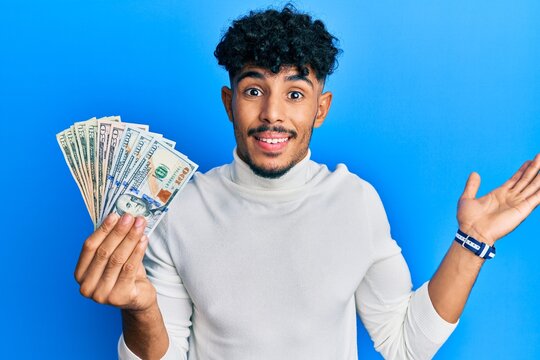 Young arab handsome man holding dollars celebrating achievement with happy smile and winner expression with raised hand