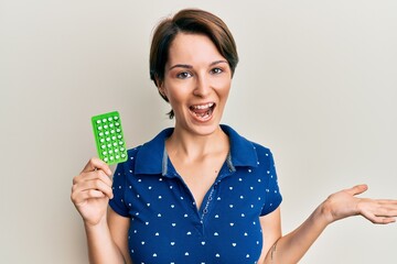 Young brunette woman with short hair holding birth control pills celebrating achievement with happy smile and winner expression with raised hand