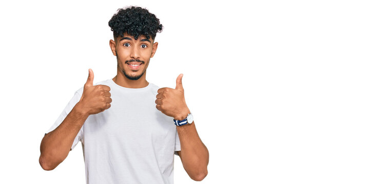 Young arab man wearing casual white t shirt success sign doing positive gesture with hand, thumbs up smiling and happy. cheerful expression and winner gesture.