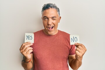 Handsome middle age mature man holding yes and no reminder winking looking at the camera with sexy...