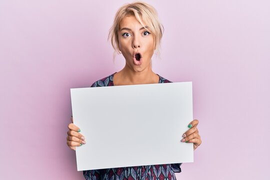 Young blonde girl holding blank empty banner scared and amazed with open mouth for surprise, disbelief face