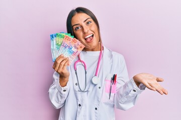 Beautiful hispanic woman wearing medical uniform holding swiss franc banknotes celebrating achievement with happy smile and winner expression with raised hand