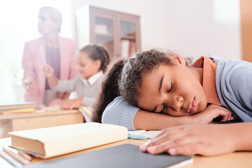 Exhausted girl keeping her head on desk on background of classmate and teacher