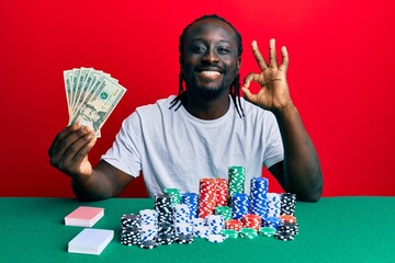 Handsome young black man playing poker holding 20 dollars banknotes doing ok sign with fingers, smiling friendly gesturing excellent symbol