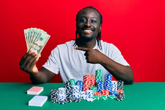 Handsome young black man playing poker holding 20 dollars banknotes smiling happy pointing with hand and finger