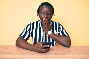 Young african american man with braids using smartphone sitting on the table covering mouth with...