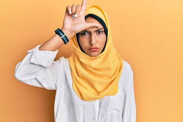 Young brunette arab woman wearing traditional islamic hijab scarf making fun of people with fingers on forehead doing loser gesture mocking and insulting.