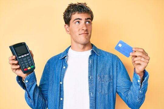 Young handsome man holding credit card and dataphone smiling looking to the side and staring away thinking.