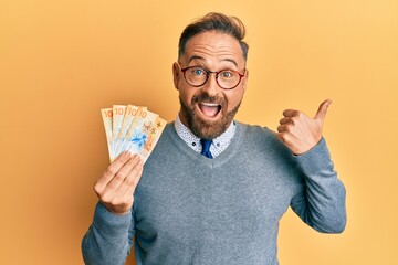 Handsome middle age man holding swiss franc banknotes pointing thumb up to the side smiling happy with open mouth