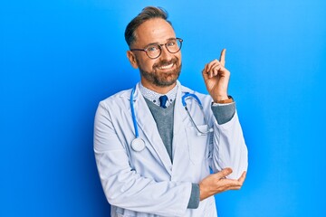 Handsome middle age man wearing doctor uniform and stethoscope with a big smile on face, pointing...