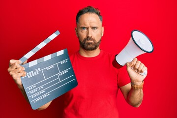 Handsome middle age man holding video film clapboard and megaphone skeptic and nervous, frowning...