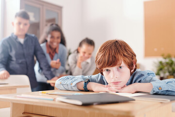 Offended schoolboy keeping chin on hands while classmates mocking at him
